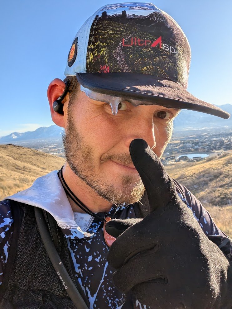 Miles Victors on a winter trail run, ice hanging from his cap brim, with mountains and a valley behind him